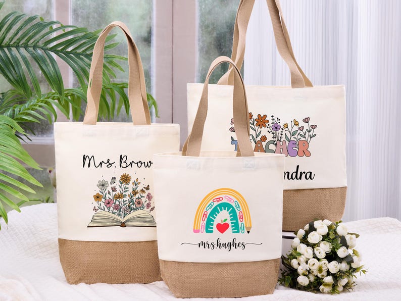 May include: Three canvas tote bags with beige handles and a burlap bottom. One bag features the text "Mrs. Brow" above an open book with flowers. Another says "Teacher" with floral accents. The third says "mrs.hughes" with a rainbow.
