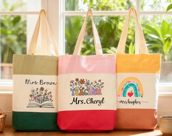 May include: Three canvas tote bags with cream handles and a two-tone design. The bags feature different graphics and text, including floral designs, a rainbow, and the names "Mrs. Brown", "Mrs. Cheryl", and "Mrs. Hughes". The bags are in shades of green, pink, and yellow.