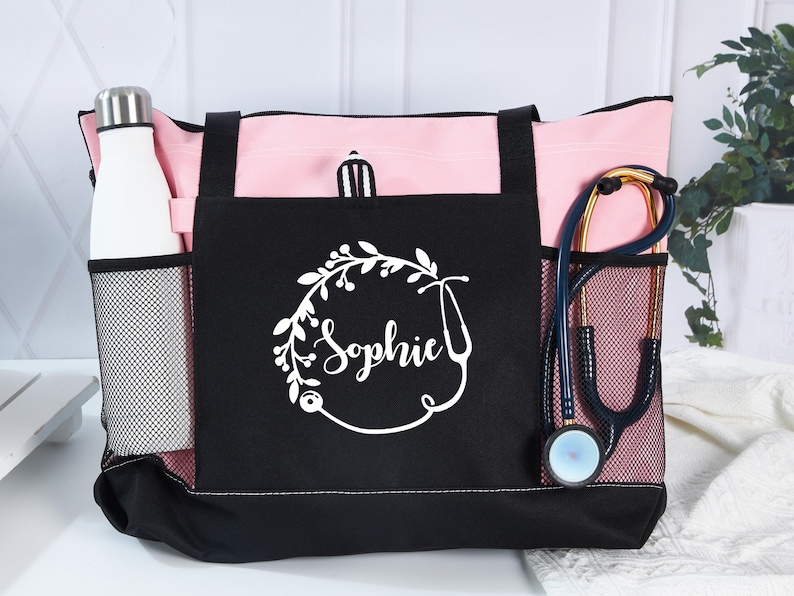 May include: Pink and black tote bag with a white floral design and the name "Sophie". The bag has a mesh side pocket with a white water bottle and another with a stethoscope. The bag is designed for medical professionals.