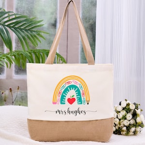 May include: Cream-colored tote bag with a burlap bottom and tan handles. The bag features a colorful rainbow design with school supplies and a red heart. The text "mrs.hughes" is written in cursive below the rainbow.