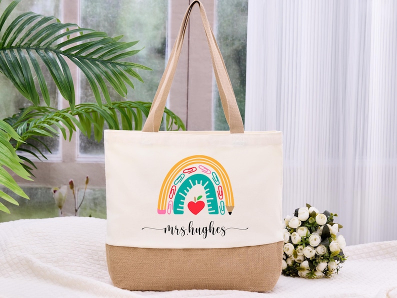 May include: Cream-colored tote bag with a burlap bottom and tan handles. The bag features a colorful rainbow design with school supplies and a red heart. The text "mrs.hughes" is written in cursive below the rainbow.