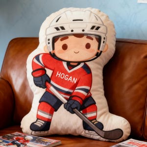 Puede incluir: Un cojín con temática de hockey con forma de jugador. El cojín presenta una ilustración de dibujos animados de un jugador de hockey con una camiseta roja con el nombre "Hogan", con un casco blanco y un palo de hockey.