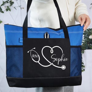 May include: A blue and black tote bag with a white stethoscope design and the name "Sophie". The bag has a blue zippered top, black handles, and mesh side pockets. The design includes the letters "RN".