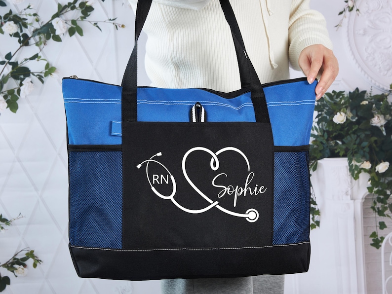 May include: A blue and black tote bag with a white stethoscope design and the name "Sophie". The bag has a blue zippered top, black handles, and mesh side pockets. The design includes the letters "RN".