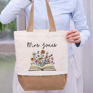 May include: Cream-colored tote bag with a floral design and the name "Mrs. Jones" in black script. The design features an open book with colorful flowers and butterflies. The bottom of the bag is a natural burlap color, and it has tan handles.