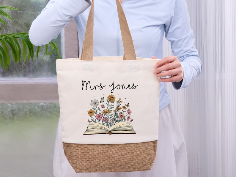 May include: Cream-colored tote bag with a floral design and the name "Mrs. Jones" in black script. The design features an open book with colorful flowers and butterflies. The bottom of the bag is a natural burlap color, and it has tan handles.