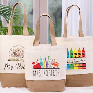 May include: Three canvas tote bags with tan handles and a burlap bottom. The bags feature teacher-themed designs, including an open book with flowers, school supplies, and a row of colorful crayons. The text "Mrs. Roberts" and "Mrs. Sandra" are visible.