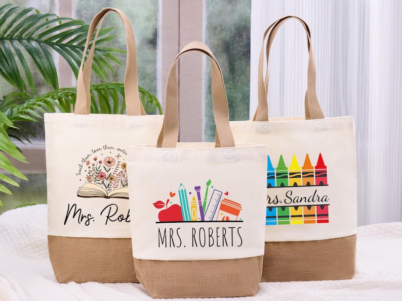 May include: Three canvas tote bags with tan handles and a burlap bottom. The bags feature teacher-themed designs, including an open book with flowers, school supplies, and a row of colorful crayons. The text "Mrs. Roberts" and "Mrs. Sandra" are visible.