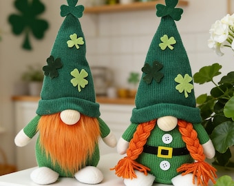 May include: Two St. Patrick's Day gnomes. Each gnome has a green knit hat with shamrock accents, a white nose, and a green body. One gnome has an orange beard, and the other has orange braids. They are sitting on a white surface.
