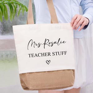 May include: Cream-colored tote bag with a tan burlap bottom and matching handles. The bag features the text "Mrs. Rosalie TEACHER STUFF" in black script and a heart symbol. A practical and stylish accessory for teachers.