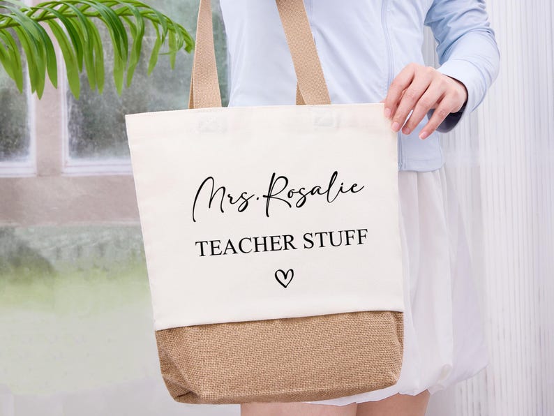 May include: Cream-colored tote bag with a tan burlap bottom and matching handles. The bag features the text "Mrs. Rosalie TEACHER STUFF" in black script and a heart symbol. A practical and stylish accessory for teachers.