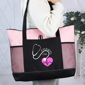 May include: A black and pink tote bag with a stethoscope and heart design. The bag has a zippered top, mesh side pockets, and black handles. The text "CHERYL CNA" is printed on the front.