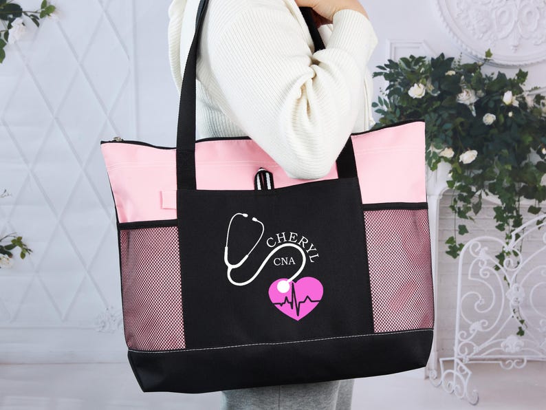 May include: A black and pink tote bag with a stethoscope and heart design. The bag has a zippered top, mesh side pockets, and black handles. The text "CHERYL CNA" is printed on the front.