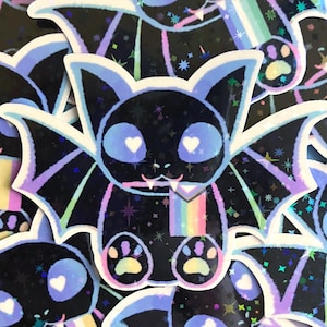 Könnte beinhalten: Eine schwarze Fledermaus mit einer Regenbogen-Pride-Flagge, blauen Augen mit weißen Herzen und Regenbogen-Pfotenabdrücken. Die Fledermaus befindet sich auf einem schwarzen Hintergrund mit Glitzer und Sternen.