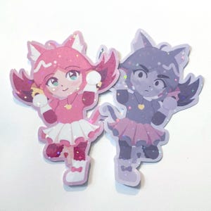 May include: Two holographic stickers featuring anime-style cat characters. The left sticker shows a pink cat character with a white skirt and heart necklace. The right sticker shows a purple cat character with a pink skirt and heart necklace.