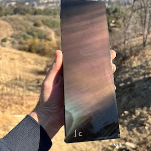May include: A dark iridescent slab of obsidian with a slight sheen. The slab is approximately 12 inches long and 4 inches wide. The text "1C" is written on the bottom right corner of the slab.