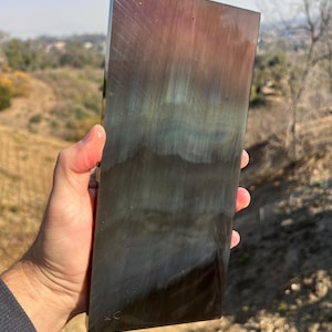 May include: A large, rectangular piece of iridescent glass with a dark green, blue, and purple color scheme. The glass has a smooth, polished surface and is held in a hand.