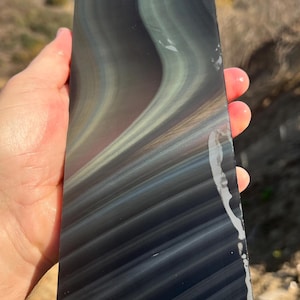 May include: A large, rectangular piece of obsidian with iridescent rainbow colors. The obsidian has a smooth, polished surface and is held in a hand.