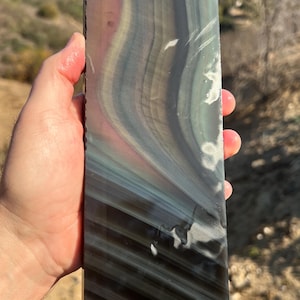 May include: A large, rectangular slab of iridescent obsidian with swirling patterns of black, gray, green, and purple. The obsidian is smooth and polished, and it has a reflective surface.