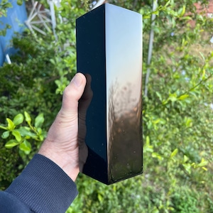 May include: A tall, rectangular black vase with a glossy finish. The vase is held against a backdrop of green foliage and a blue structure. The vase's surface reflects the surrounding environment, creating a gradient effect.