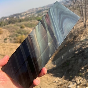 May include: A large, iridescent obsidian slab with a dark, black base and streaks of blue, green, and purple. The slab is smooth and polished, and the colors are vibrant and eye-catching.