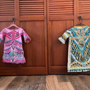 May include: Two colorful dresses hang on wooden doors. The dress on the left is pink with blue and black geometric patterns, fringe, and floral accents. The dress on the right is blue, yellow, and green with intricate designs and a ruffled hem.
