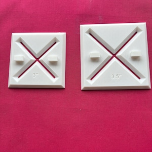 May include: Two white square quilting templates with an X design and small rectangular tabs. The smaller template is 3 inches, and the larger is 3.5 inches. The templates are on a pink background.