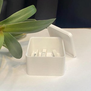 May include: A white square box with a lid. The box is open and has a few small white objects inside.