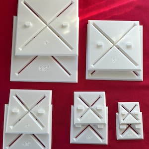 May include: A collection of white, square quilting templates in various sizes, ranging from 2.5 inches to 6.5 inches. Each template features an X-shaped design with diagonal lines and measurement labels in inches. The templates are stacked on a red surface.