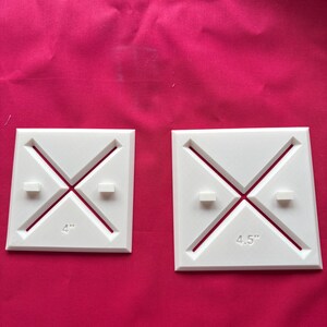 Könnte beinhalten: Zwei weiße quadratische Quiltwerkzeuge mit einem Kreuzdesign und abgewinkelten Linien. Ein Werkzeug ist mit "4" und das andere mit "4,5" Zoll (ca. 11,4 cm) beschriftet. Die Werkzeuge befinden sich auf einem rosa Hintergrund.