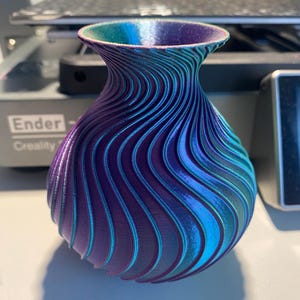 Puede incluir: Un jarrón impreso en 3D con un acabado azul y morado iridiscente en espiral. El jarrón tiene una base ancha y un cuello estrecho.