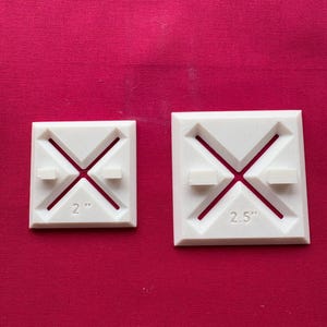 Pode incluir: Dois objetos quadrados brancos com um design em X e um pequeno bloco retangular em cada lado. O objeto menor está rotulado como "2" e o maior como "2.5". Os objetos estão em um fundo vermelho.