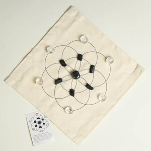 May include: A white canvas bag with a black geometric design and clear and black crystals arranged in a circle. The bag includes a small card with the text "Protection and Cleansing Crystal Grid Set".