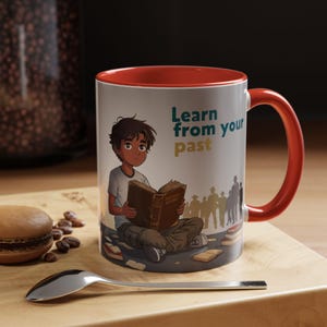 May include: A white ceramic mug with a red interior and handle. The mug features an illustration of a young person reading a book with the text "Learn from your past". The mug is on a wooden surface.