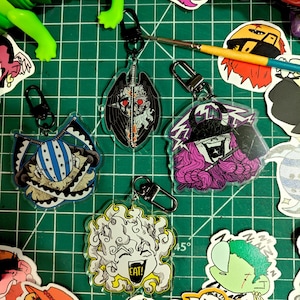 May include: A collection of acrylic keychains with various cartoon character designs. The keychains feature black metal clasps and depict characters with bold colors and intricate details. The background is a green cutting mat with other stickers and art supplies.
