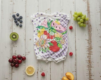 May include: White baby romper with a fruit-themed design. The romper features a floral and fruit print, including watermelon, cherries, and flowers. The top has purple polka dots and a checkered pattern. Fresh fruit like blueberries, grapes, and raspberries are arranged around the romper.