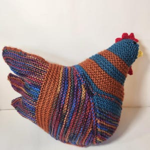 May include: A knitted chicken-shaped pillow in shades of blue, orange, and red. The body features horizontal stripes, while the head is blue with a red comb and yellow beak. The wings are multi-colored.