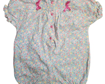 May include: A light blue baby romper with a floral pattern in pink, yellow, and blue. The romper has short, ruffled sleeves, pink bows, and pink buttons down the front. The bottom hem is also ruffled.