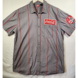 May include: Gray short-sleeve button-up shirt with red vertical stripes. Features a Coca-Cola logo patch on the chest pocket and a Coca-Cola logo patch on the sleeve. The shirt has a collared neckline and a button-down front.