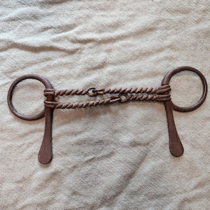 May include: A dark brown metal horse bit featuring two circular rings connected by a double-jointed mouthpiece. The mouthpiece consists of two twisted metal bars that link together in the centre, with flat, downward-extending shanks on each side.