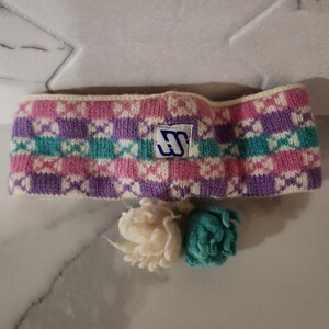 May include: A knitted headband in a geometric pattern of pink, purple, teal, and white. The headband features a small label with a logo. Two small pom-poms, one cream and one teal, are below the headband.