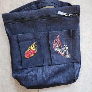 May include: Dark blue denim tote bag with a zip closure and two front pockets. The pockets feature embroidered patches of a motorcycle and a flaming die. The bag has a handle for carrying.