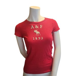 May include: A short-sleeved red t-shirt featuring the white embroidered text A & F, a white embroidered moose graphic, and the white embroidered number 1892 centred on the chest. The garment has a crew neckline and a fitted silhouette.