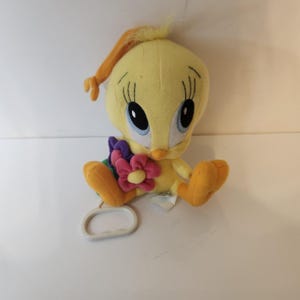 May include: A yellow plush Tweety Bird character with large blue eyes, long eyelashes, and orange feet. The plush toy holds a cluster of purple and pink fabric flowers in its lap and features a white plastic pull-ring handle attached at the base.