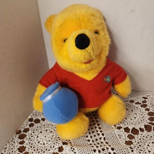 Peut inclure: Une peluche Winnie l'ourson, avec une fourrure jaune, un t-shirt rouge à col en V et un pot de miel bleu. Le jouet a un nez noir et une expression souriante. Le t-shirt porte le mot "Pooh".