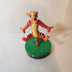 May include: A plastic figurine of a tiger character with orange fur, black stripes, and a cream-colored belly. The character is posed with arms outstretched and legs bent, mounted on a round green base with small blue flower details.