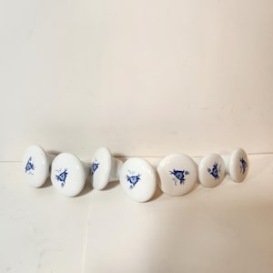 Op de afbeelding: Een verzameling van acht witte keramische kastknoppen met een blauw bloemmotief. De knoppen variëren in grootte en vorm, met een ronde of ovale vorm. Het blauwe bloemmotief is gecentreerd op elke knop.
