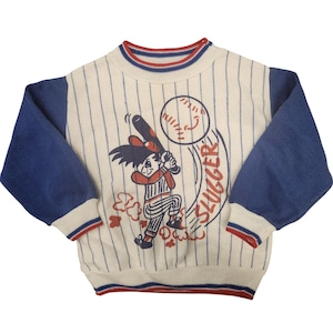 May include: Cream-coloured baseball sweater with blue sleeves and red trim. The front features a cartoon illustration of a batter swinging at a baseball, with the word "SLUGGER" in red. Vertical blue stripes run down the front.