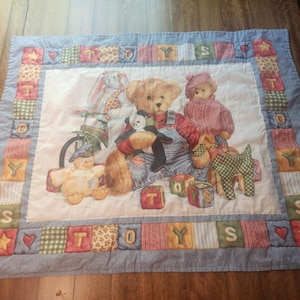 Op de afbeelding: Een gewatteerd stoffen wandkleed met een centrale illustratie van teddyberen, een konijn en een eend met speelgoed. De rand bestaat uit patchworkvierkanten in blauw, groen, rood en geel, met de letters T, O, Y, S en hartvormige motieven.