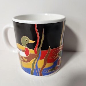 以下が含まれることがあります： 白いハンドルと縁が付いたセラミックコーヒーマグ。マグカップは、黒い背景に、水中でアシとともに泳ぐアヒルのカラフルなイラストが特徴です。アヒルは緑、黄色、赤、茶色の色合いです。